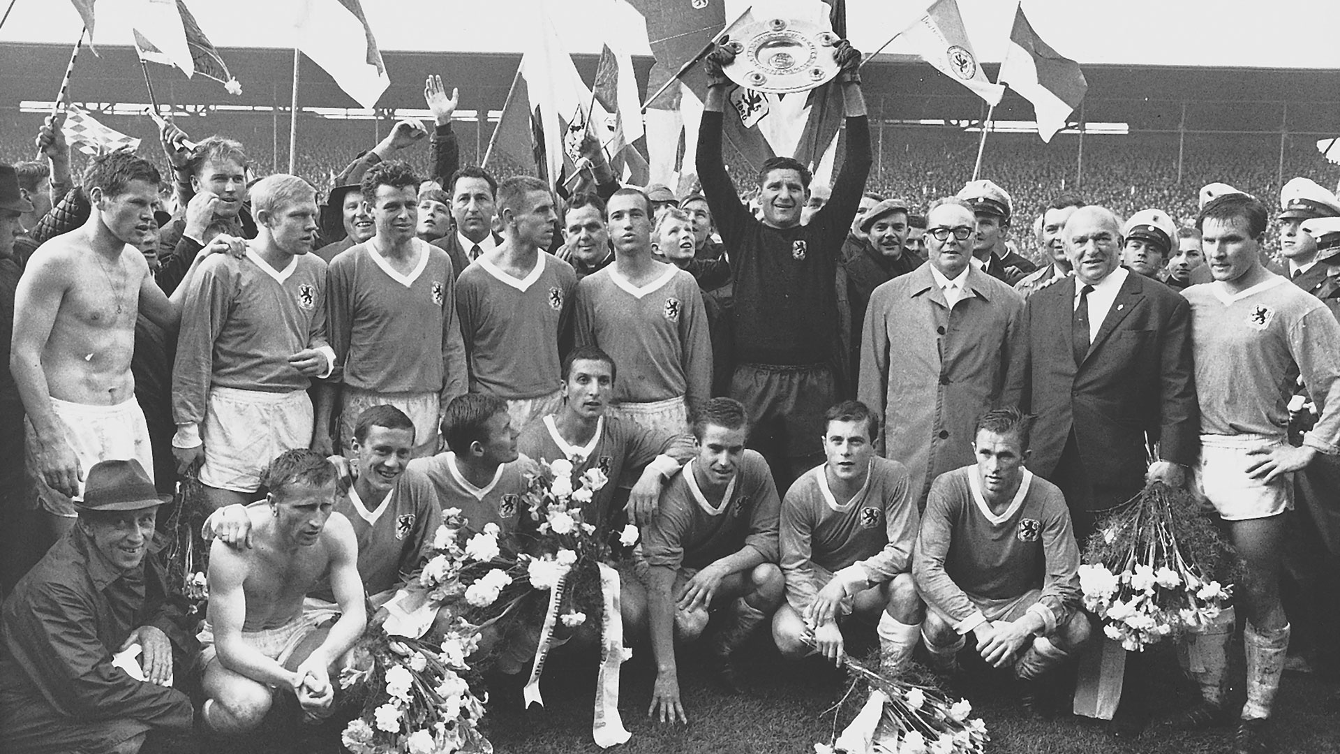 Schwarzweissaufnahme von 1966: die Mannschaft des TSV 1860 München steht mit der Meisterschale im Stadion, dahinter wehen Fahnen